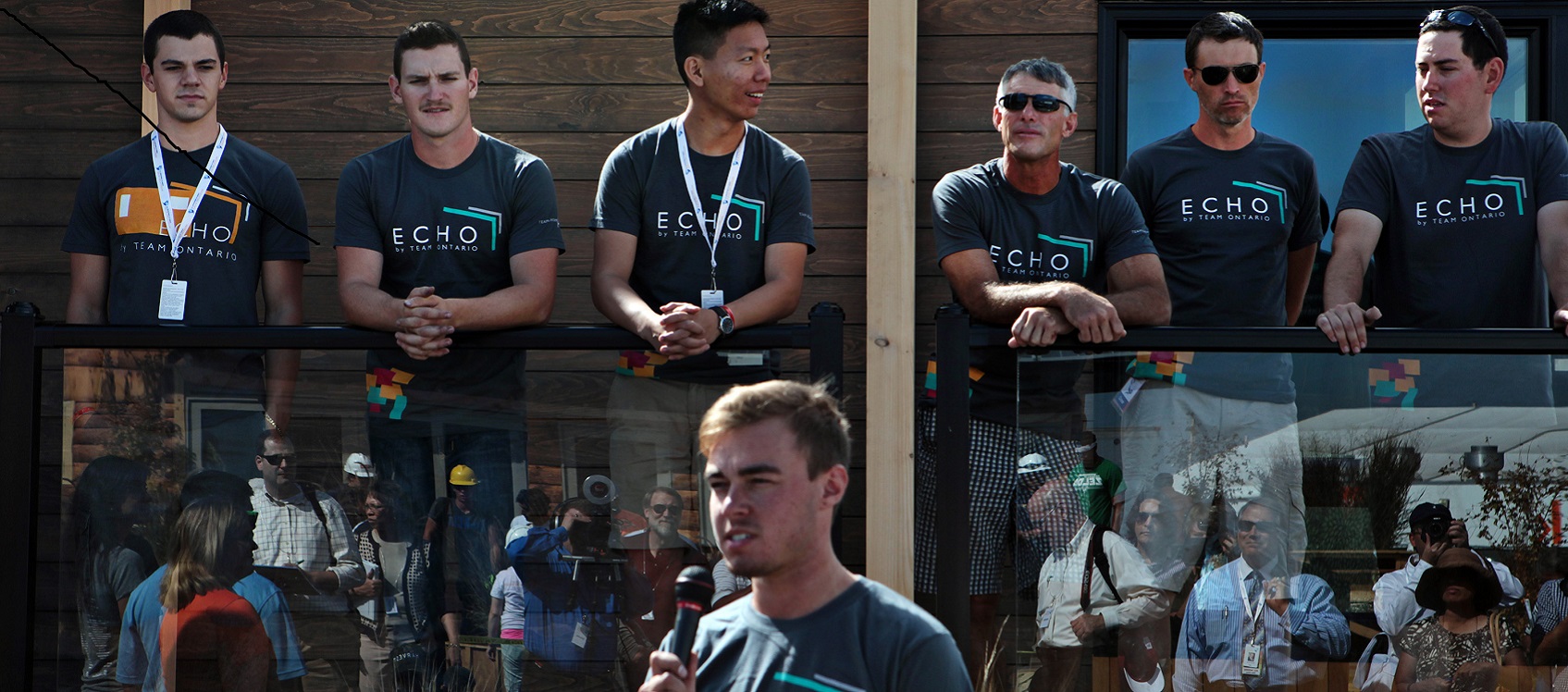 A team of college students stands on the porch of their Solar Decathlon home while one student speaks into a microphone.