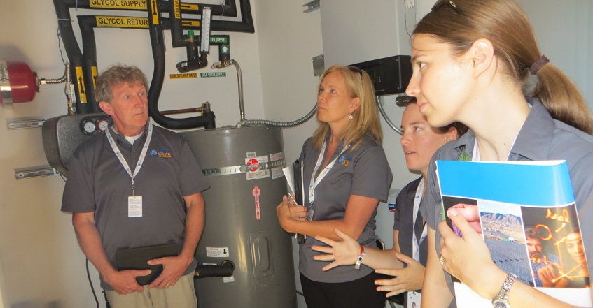 Photo of a man and two women who served on the U.S. Department of Energy Solar Decathlon 2015 Engineering contest jury are standing in an equipment room with solar thermal and solar electric equipment behind them. They are wearing gray uniform shirts and listening to a female student from the Cal Poly team, who is presenting the team’s engineering presentation to the jury.