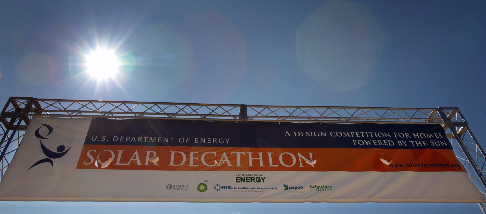 Photo of a banner sign, outdoors, with the sun shining behind it. The blue, orange and white banner welcomed visitors to entrance of the Solar Decathlon 2009 solar village in Washington, D.C. and features event sponsor logos.