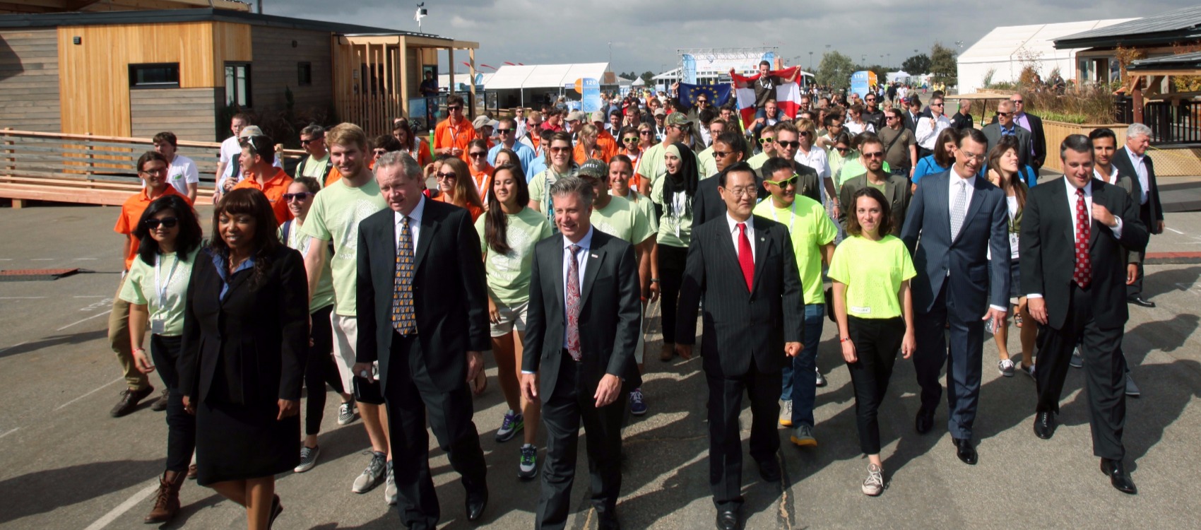 Photo of a group of sponsor representatives-men and women in business attire—leading a larger group of young men and women—all student team members of the Solar Decathlon 2013 teams—as they enter the Solar Decathlon competition village in Irvine, California. Team houses, white event tents, and signs are visible in the background.