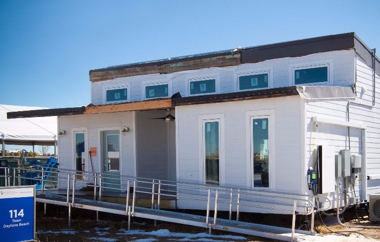 Photo of the exterior of a white house with a ramp approaching a pale blue front door, multiple vertical windows and clerestory windows above. There is a sign indicating this the Team Daytona Beach house for the U.S. Department of Energy Solar Decathlon 2017.
