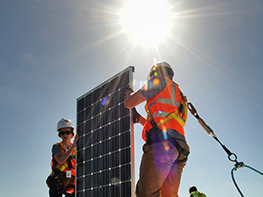 Photo of a woman and man, both wearing yellow hard hats and orange safety vests carrying a photovoltaic panel (solar electric panel) on the roof of a Solar Decathlon house. The sun is shining brightly in the sky behind them. They are both clipped into safety harnesses appropriate for working at heights.