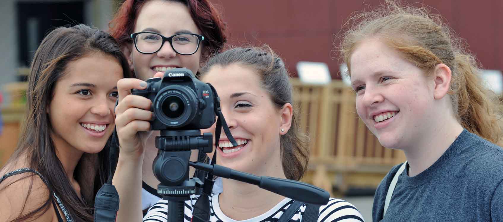 Photo of four young women, all U.S. Department of Energy Solar Decathlon 2015 competitors, smiling and looking at the display on the back of a camera mounted on a tripod.