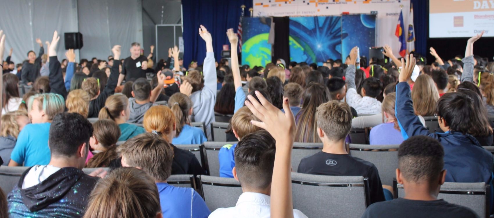 Photo of backs of middle school boys and girls sitting in chairs and raising their hands to answer questions during an education session inside a white tent at Solar Decathlon 2017. The students are facing a stage and a projection screen.