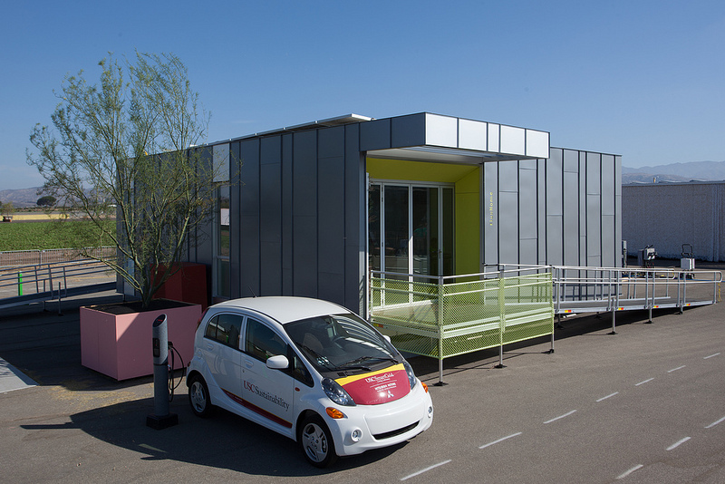 Photo of an electric vehicle parked next to a charging station beside a Solar Decathlon competition house. 