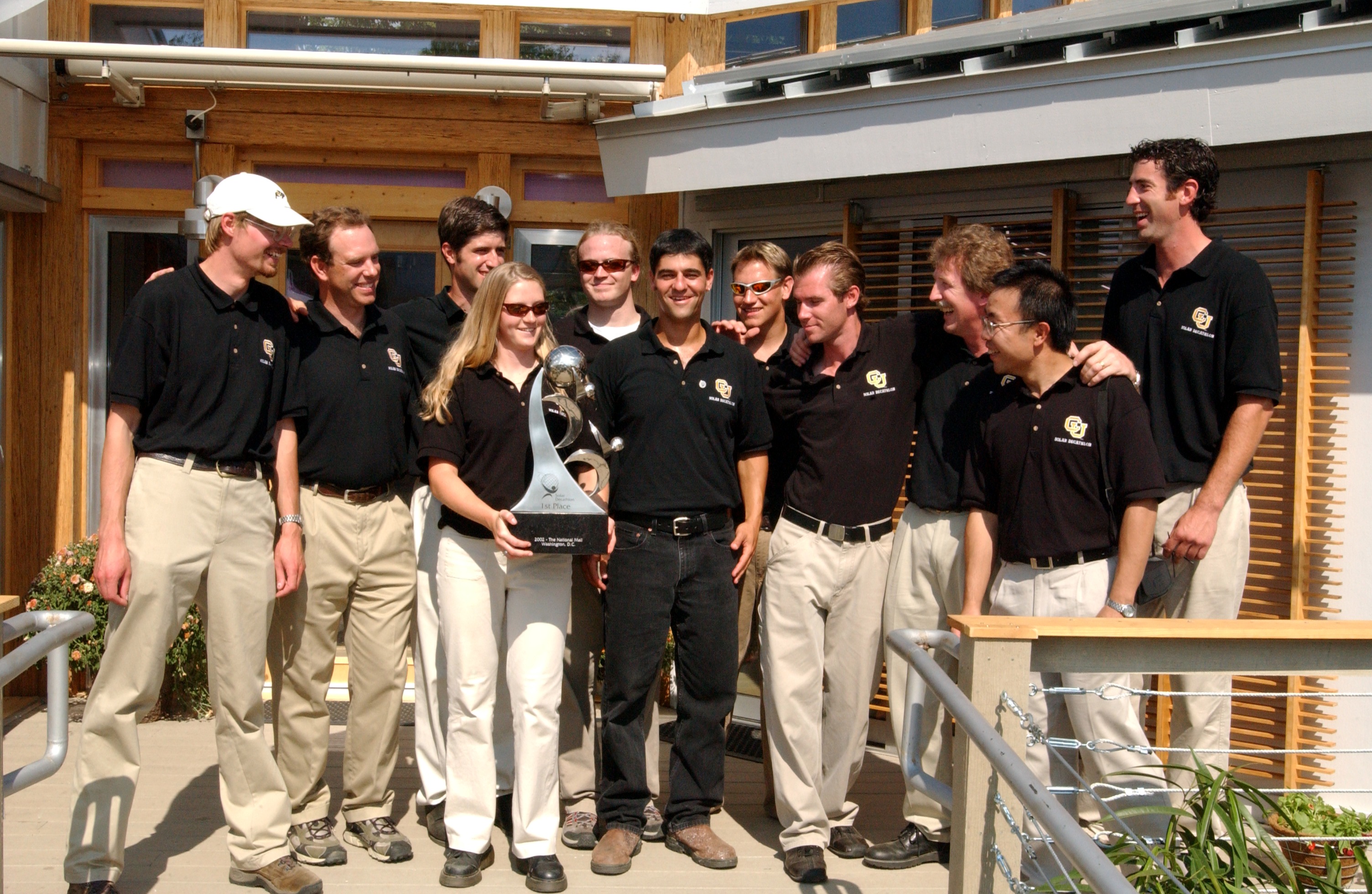 Photo of a group of men standing around a woman who is holding a large trophy.