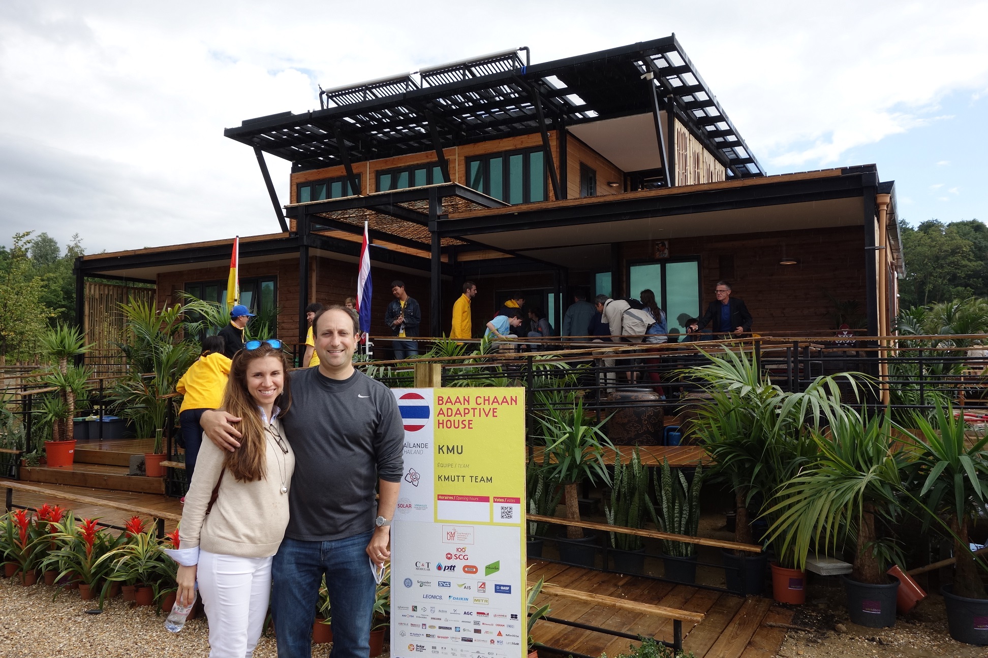 Photo of a man with his arm around a woman in front of a modern-looking wooden house.
