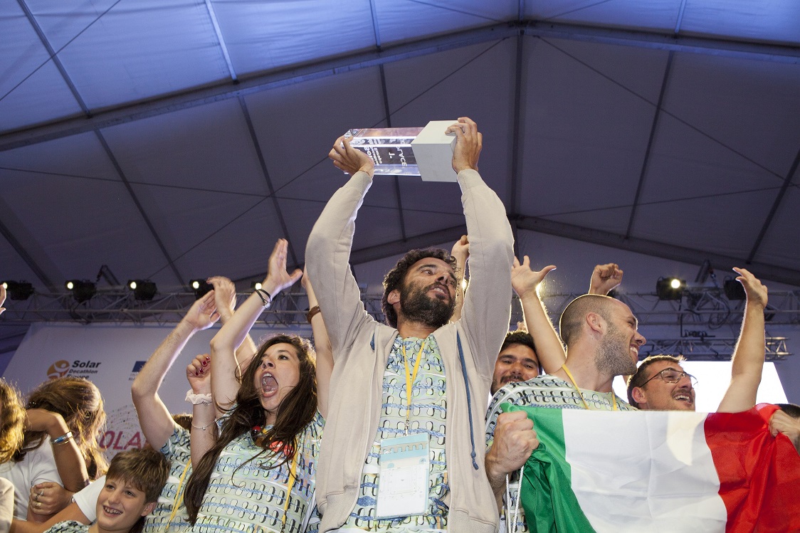 Photo of a group of cheering people holding an Italian flag and a first-place trophy.