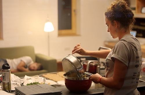Photo of a woman cooking at a counter. In the background, a man sleeps on a couch.