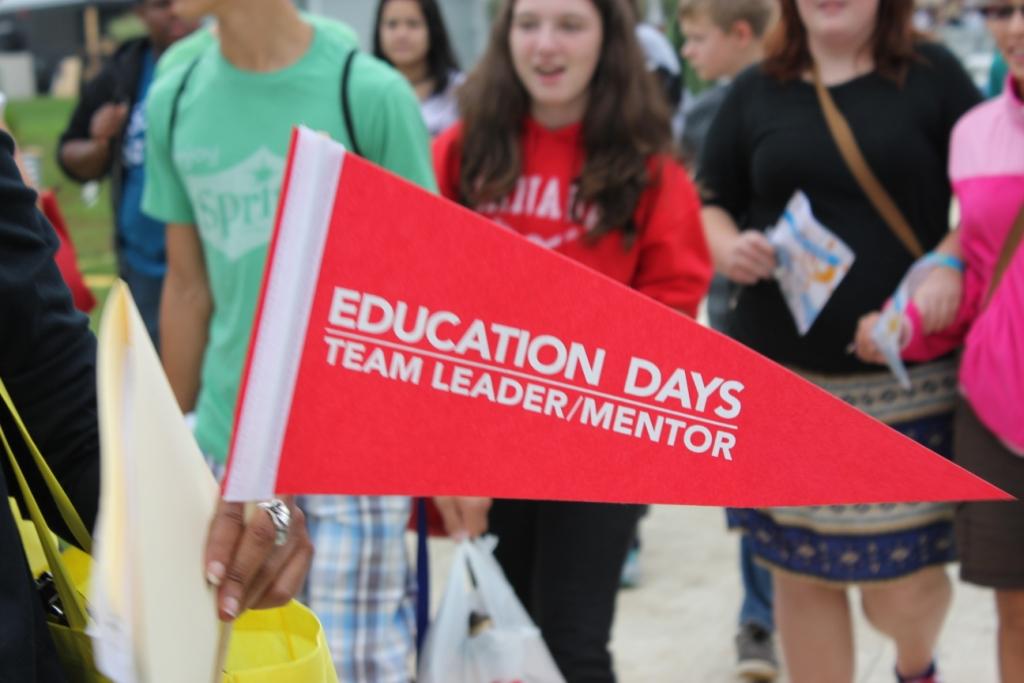 Photo of a red flag that says “Education Days Team Leader/Mentor.” In the background is a group of students. 