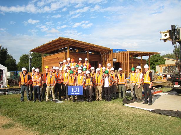 Photo of a group of people wearing hard hats, safety vests, and safety glasses standing in front of a house. A sign in front reads “101: New Zealand.”