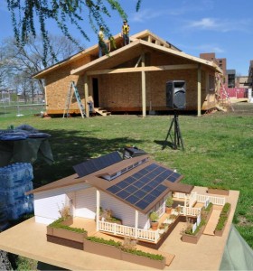 Photo of a model of a house on a table beneath a tree. In the background is the full house under construction. 