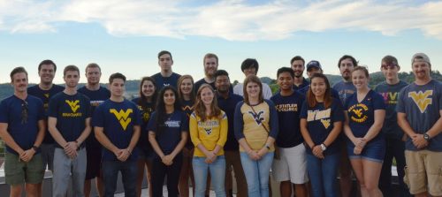 Photo of the Solar Decathlon 2017 team from West Virginia University; they are a group of about 20 students, men and women, smiling and wearing various designs of blue and gold WVU t-shirts. A skyline featuring a ridge of Appalachian Mountains is in the background.