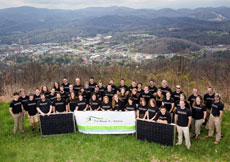 Photo of a group of people in matching shirts standing on a hill overlooking a campus. In front, the team holds two PV panels and a sign that says "The Solar Homestead: Appalachian State University. Appalachian State Solar Decathlon Headquarters."