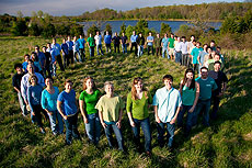 Photo of a group of people standing in a circle in a field. In the background is a body of water.