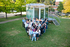 Photo of a group of people standing in a V shape in front of a glass building with PV panels.