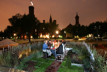 Photo of people eating dinner by candlelight in a garden.