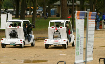 Photo of two people, one with a camera boom, driving an electric vehicle next to another electric vehicle with a driver and passenger.