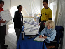 Photo of a blond-haired woman sitting at a table that holds a large pile of folded towels. Two young men and one young woman are also visible in the photo.