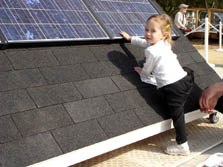 Photo of a 2-year-girl reaching out to touch a solar panel that is displayed on a section of a ground-based roof with shingles.