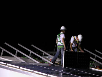 Photo of two young men on a roof, wearing protective gear and installing a solar panel.
