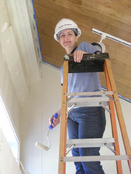 Photo of a smiling student in a hard hat, standing on a ladder and holding a paint roller.