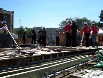 Photo of ten to twelve people working on top of a partially completed deck, with metal support work visible and tools lying about.