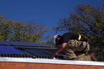 Photo of a man in a hard hat on the roof of a solar home, kneeling down and lifting the end of a glass tube.
