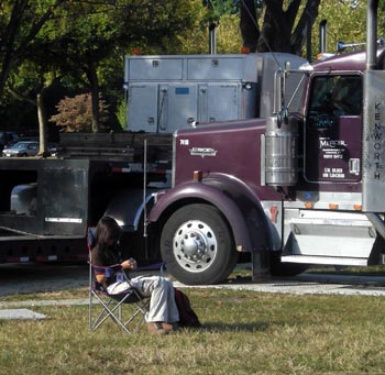 Photo of a woman sitting in a folding chair between two tractor-trailer trucks, holding a mobile telephone.