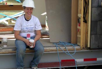 Photo of a woman in a hard hat holding a cup of coffee, sitting on the edge of an opening in a partially dismantled solar home.