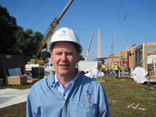 Photo of Richard King, who is wearing a hard hat and standing in front of a construction site. Multiple houses, cranes, trucks, and roll-offs, as well as the Washington Monument, can be seen in the background.