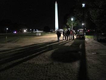 Photo of a group of people standing in the dark beneath a streetlight on the National Mall. The lighted Washington Monument is in the background.