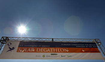 Photo of banner suspended by a metal frame. The banner says "U.S. Department of Energy Solar Decathlon: A Design Competition for Homes Powered by the Sun." Above the banner and the frame is the sun.