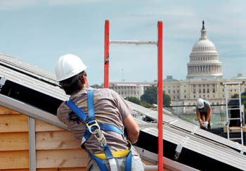 Photo of man wearing a hard hat and safety harness standing on a ladder leaning against the roof of a house. In the background, a woman works atop a neighboring roof. The U.S. Capitol is seen further away.