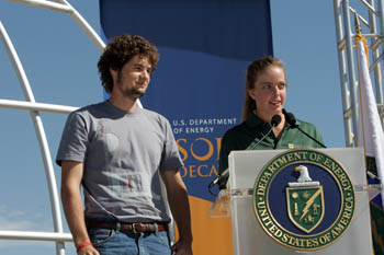 Photo of a young woman speaking from behind a podium with the U.S. Department of Energy seal. A young man stands next to her.