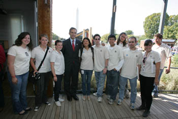 Photo of Michael Arcuri, wearing a suit, standing among a group of students in matching white shirts on the porch of the Cornell University house.