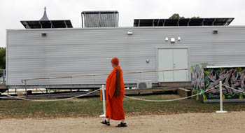 Photo of a man in orange hats and robe walking past a long, rectangular, metal-clad structure.