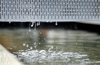 Photo of water drops falling from a perforated metal sheet and splashing into a puddle below.