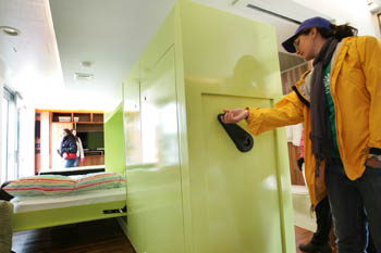 Photo of a woman turning a lever on a wall unit in the middle of a house. On one side of the unit, a bed protrudes.