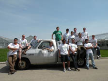 Photo of fourteen team members, who are standing around and in the bed of a white pickup truck. The truck is parked in front of the University Greenhouses.