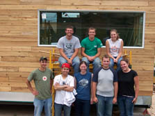 Photo of eight students sitting on and standing around scaffolding in front of their project house.