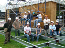 Photo of 12 Puerto Rico team members in front of their unfinished house. Some team members sit on or stand inside a metal grid. The others sit on scaffolding.
