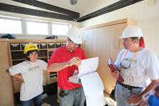 Photo of two men and one woman reviewing documents. All wear hardhats and stand inside a partially finished room.