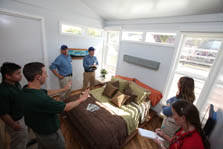 Photo of a group of people in a small bedroom. Two onlookers take notes as two student decathletes speak to them. 
