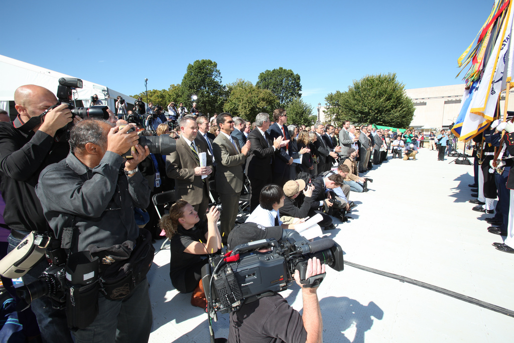 Photo of members of the news media, men and women, holding cameras and video cameras, standing outside in the sunshine in front of a stage decorated with flags.