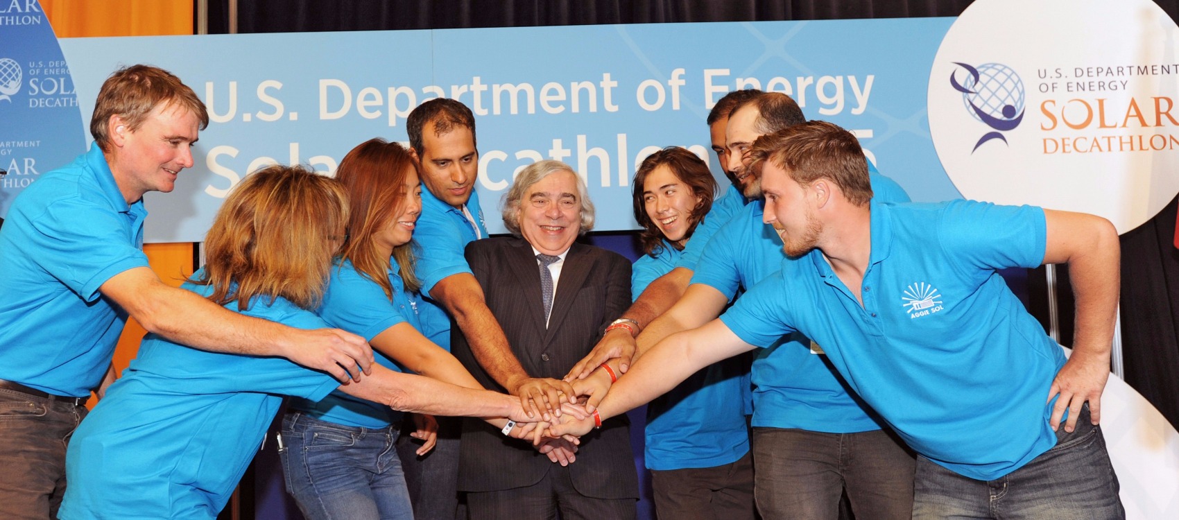 Photo of a man with shoulder-length white hair, Secretary of Energy, Dr. Ernest Moniz, wearing a suit and tie surrounded by seven men and women in blue shirts, all student team members of the Solar Decathlon 2015 team from the University of California, Davis. Each person has one hand on top of the others’ hands in preparation for a cheer of team spirit.