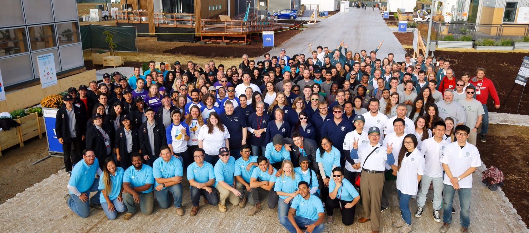 Photo of a large group of young men and women, all members of the teams of the U.S. Department of Energy Solar Decathlon 2017, standing on a temporary walkway between rows of full-sized solar powered houses.