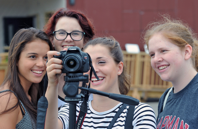Photo of four young women, all U.S. Department of Energy Solar Decathlon 2015 competitors, smiling and looking at the display on the back of a camera mounted on a tripod.