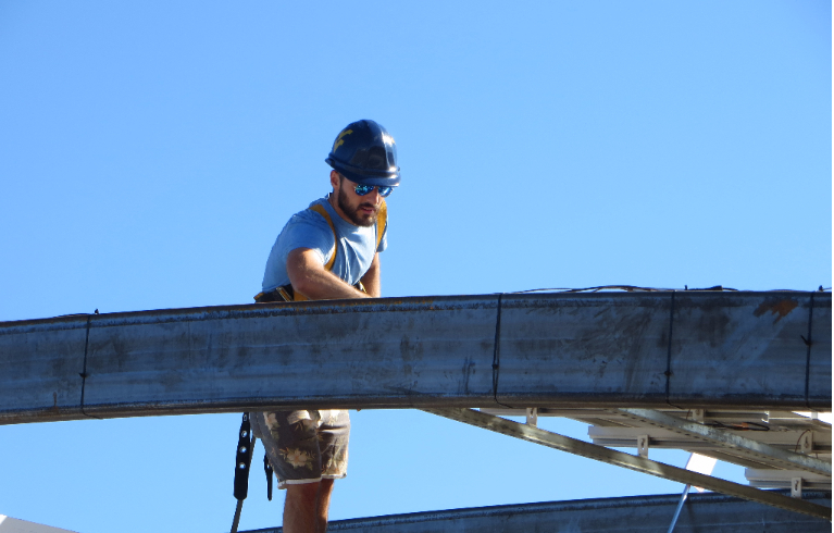 Photo of a young man, a competitor in the U.S. Department of Energy Solar Decathlon 2015, wearing a blue hard hat and blue reflective sunglasses on top of a roof. Above the roof are steel girders supporting photovoltaic solar electric panels. The man is cleaning the panels. Behind him is a clear blue sky.