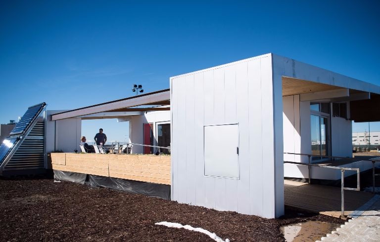 Photo of the exterior of a modern house with an extended roof over a large front deck. The black glass tubes of an evacuated tube solar collector are visible to the side of the deck. This is the Sinatra Living house by the U.S. Department of Energy Solar Decathlon 2017 team from the University of Nevada, Las Vegas.